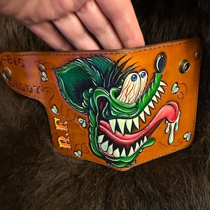 Può includere: Un portafoglio in pelle color cuoio con il disegno dipinto a mano di un topo verde in stile cartoon, con occhi sporgenti, denti affilati e una lunga lingua rosa. Il testo "BIG DADDY" e "R.F." è impresso a sinistra. Piccole mosche sono dipinte intorno.