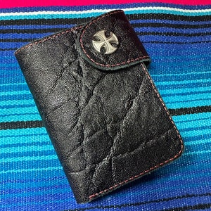 May include: A black leather wallet with a cross-shaped metal accent. The wallet has a textured surface and a red stitching detail.
