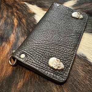 Op de afbeelding: Zwarte leren portemonnee met een getextureerd oppervlak en zilveren schedelaccenten. De portemonnee heeft een metalen ring en is vastgezet met metalen klinknagels. De rechthoekige portemonnee lijkt gesloten.