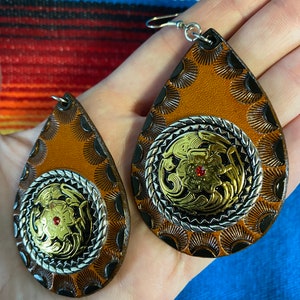 Op de afbeelding: Een paar druppelvormige oorbellen met een western thema. De oorbellen hebben een bruine leren basis met zilver- en goudkleurige metalen details. Een kleine rode edelsteen is in het midden van het goudkleurige metalen ontwerp.