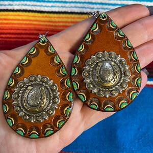 Op de afbeelding: Een paar bruinleren druppelvormige oorbellen met zilverkleurige metalen versieringen. Elke oorbel heeft een hoefijzer- en bloemmotief in het midden, omgeven door een decoratieve rand met turquoise en gouden accenten. De oorbellen zijn ontworpen voor doorboorde oren.