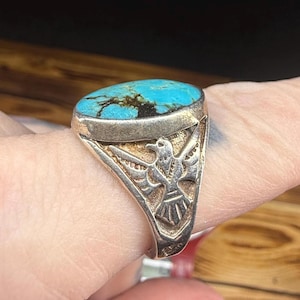 May include: A silver ring featuring a large oval turquoise stone. The turquoise is a bright blue with dark brown markings. The ring has an eagle design on the sides.