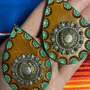 Op de afbeelding: Een paar druppelvormige oorbellen met een western thema. De oorbellen hebben een bruine lederen basis met turquoise en gele accenten. Elke oorbel heeft een zilverkleurige metalen versiering met een hoefijzerontwerp in het midden.
