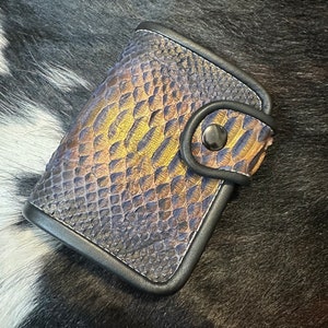 May include: A small, black leather wallet with a gold and blue snakeskin pattern. The wallet has a snap closure and a single button.