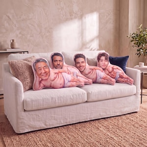 May include: Four custom-shaped throw pillows arranged on a white sofa. Each pillow features a printed illustration of a different person's head attached to a muscular torso wearing white briefs, with each figure posed in a reclining position.