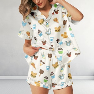 May include: A short-sleeved button-down pajama shirt and matching shorts set in off-white, featuring an all-over print of various food items shaped like mouse ears, including burgers, pizza slices, ice cream cones, sundaes, and blue cotton candy.