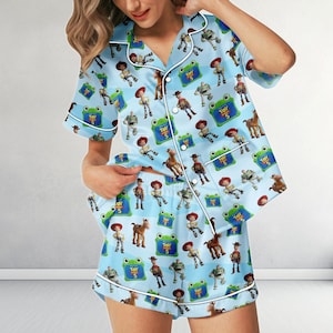 May include: A light blue pajama set consisting of a short-sleeved button-down shirt and matching shorts. The fabric features a repeating pattern of Toy Story characters including Woody, Buzz Lightyear, and green frogs with the text Toy Story 5.
