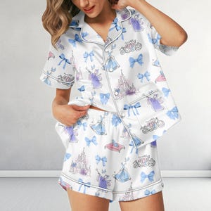 May include: White satin pajama set featuring a repeating pattern of Cinderella-themed illustrations in blue, purple, and pink. The set includes a button-down shirt with short sleeves and shorts, both trimmed with white piping.