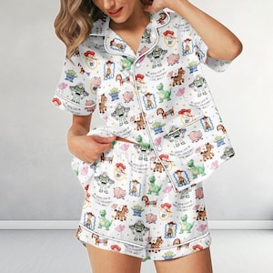 May include: A white pajama set featuring a short-sleeved button-down shirt and matching shorts. The fabric is covered in a colorful repeating pattern of cartoon characters including a cowboy, space ranger, cowgirl, pig, and green alien.