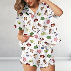May include: A white short-sleeved button-down pajama shirt and matching shorts set featuring a repeating pattern of various cartoon characters including a cowboy, a space ranger, a green alien, a dinosaur, a pig, and a cowgirl, with small yellow stars.