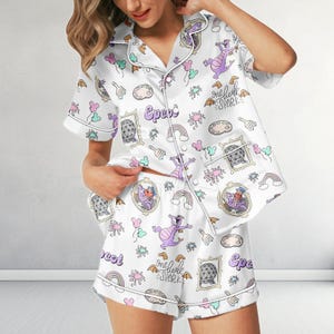 May include: White satin pyjama set with a whimsical print featuring a purple dragon, rainbows, and the word "Epcot". The set includes a button-down shirt and shorts, both trimmed with white piping.