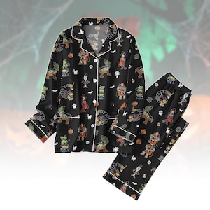 May include: A black long-sleeve pajama set featuring a repeating pattern of cartoon characters in Halloween costumes, including aliens, a cowboy, and a spork, interspersed with pumpkins, ghosts, bats, and skulls, with white piping trim.