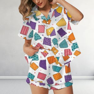 May include: A white pajama set consisting of a short-sleeved button-down shirt and matching shorts. The fabric features a colorful patchwork pattern of squares in shades of orange, purple, pink, and teal, each with distinct geometric textures.