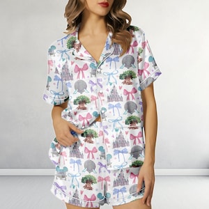 May include: White pajama set featuring a pattern of castles, bows, trees, and other graphics in pink, blue, and grey. The short-sleeved top and shorts have white trim. The set is made of a silky material.