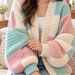 May include: A chunky knit cardigan featuring a color-blocked pattern of cream, pastel pink, and mint green squares. The garment has long sleeves with ribbed cuffs, an open front design, and a thick ribbed collar that extends down the front.