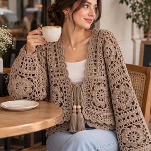 May include: A beige crochet cardigan featuring intricate floral motifs, long sleeves, and a scalloped edge. The garment includes a front tie closure adorned with two wooden beads and matching tassels, creating a textured, open-knit appearance.