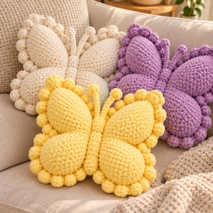 May include: Three butterfly-shaped pillows crafted from chunky yarn in yellow, purple, and cream. Each pillow features a textured, bobble-stitch border and a central body section, with two small antennae extending from the top of each butterfly shape.