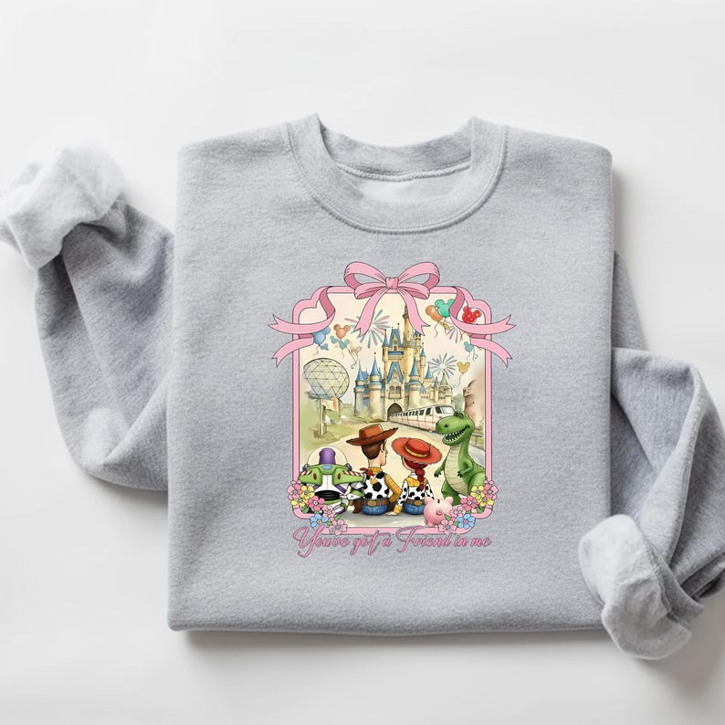 May include: A light gray sweatshirt featuring a colorful graphic of Toy Story characters in front of a castle and fireworks. The graphic includes the text "You've got a friend in me" in a script font. The sweatshirt is made of a soft material.