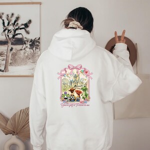 May include: White hoodie with a colorful graphic on the back featuring a castle, characters, and the text "You've got a friend in me." The graphic is framed by a pink bow and ribbon design.