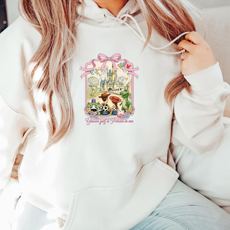 May include: Cream-colored hoodie featuring a colorful graphic of a castle, characters, and the text "You've got a Friend in me." The graphic includes a pink bow and floral accents. The hoodie has a drawstring hood.