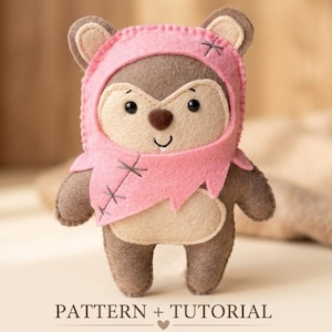 May include: A handmade felt doll in the shape of a woodland creature, wearing a pink hood and scarf. The doll has a tan face, brown eyes, and a small smile. The text "PATTERN + TUTORIAL" is at the bottom.