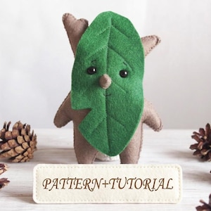 May include: A handmade felt doll resembling a tree spirit, with a green leaf face and brown body. The doll has small arms and ears, and a smiling expression. The text "PATTERN+TUTORIAL" is on a small tag in front of the doll. Pinecones are scattered around.