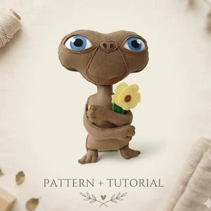 May include: A brown felt E.T. doll with large blue eyes, holding a yellow flower. The doll has stitched details and is standing on a neutral background. The text "PATTERN + TUTORIAL" is below the doll.