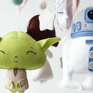 May include: A handmade baby mobile featuring felt characters from Star Wars. The mobile includes a green Yoda, a white and blue R2-D2, and other decorative elements. The characters are suspended by strings, creating a whimsical nursery decoration.