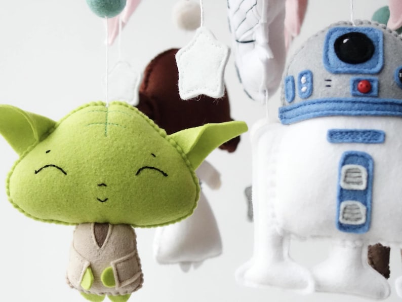 May include: A handmade baby mobile featuring felt characters from Star Wars. The mobile includes a green Yoda, a white and blue R2-D2, and other decorative elements. The characters are suspended by strings, creating a whimsical nursery decoration.
