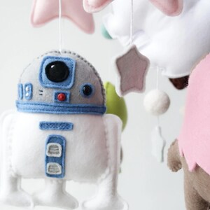 May include: A handmade baby mobile featuring felt characters and shapes. The mobile includes a white and blue R2-D2 droid, pink stars, and other colorful felt elements. The mobile is designed to hang from the ceiling.