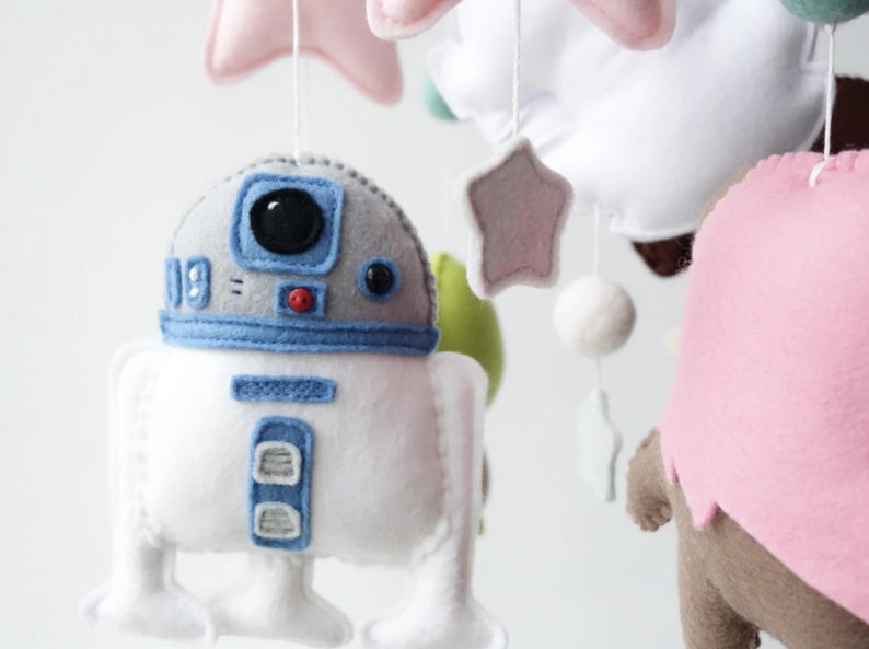 May include: A handmade baby mobile featuring felt characters and shapes. The mobile includes a white and blue R2-D2 droid, pink stars, and other colorful felt elements. The mobile is designed to hang from the ceiling.