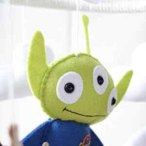 May include: A handmade felt mobile featuring a green alien character with three eyes, large ears, and a blue shirt. The alien is part of a baby mobile with white cloud shapes in the background.