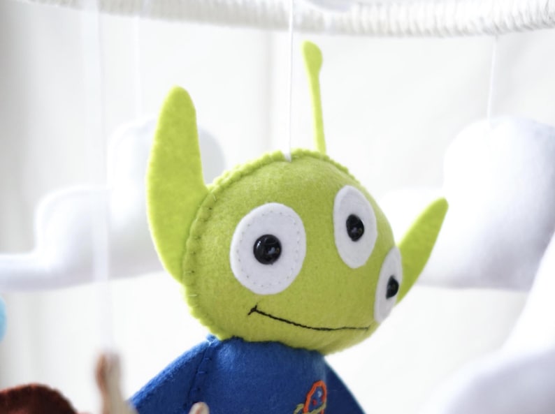 May include: A handmade felt mobile featuring a green alien character with three eyes, large ears, and a blue shirt. The alien is part of a baby mobile with white cloud shapes in the background.