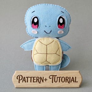 May include: A handmade plush toy of a cartoon turtle character in light blue, with a yellow shell and pink accents. The toy has large, expressive eyes and a smiling face. A wooden sign below reads "PATTERN + TUTORIAL".