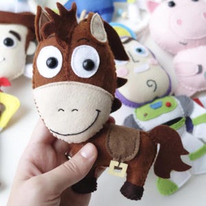 May include: Handmade felt plush toys of characters from the animated movie Toy Story. The toys include a brown horse, Woody, Buzz Lightyear, and others. The horse has a light tan muzzle and a brown saddle.
