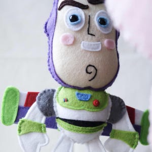 May include: A handmade felt Buzz Lightyear doll. The doll features a white face with pink cheeks, a purple helmet, and a green and white spacesuit. The wings are green, red, and white. The doll is a toy.