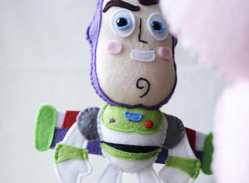 May include: A handmade felt Buzz Lightyear doll. The doll features a white face with pink cheeks, a purple helmet, and a green and white spacesuit. The wings are green, red, and white. The doll is a toy.