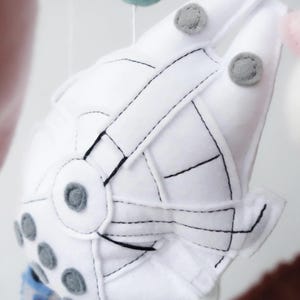 May include: A white felt mobile element shaped like a spaceship, with black stitched details and gray accents. The spaceship has a circular design with gray circles and is suspended with other colorful felt shapes.