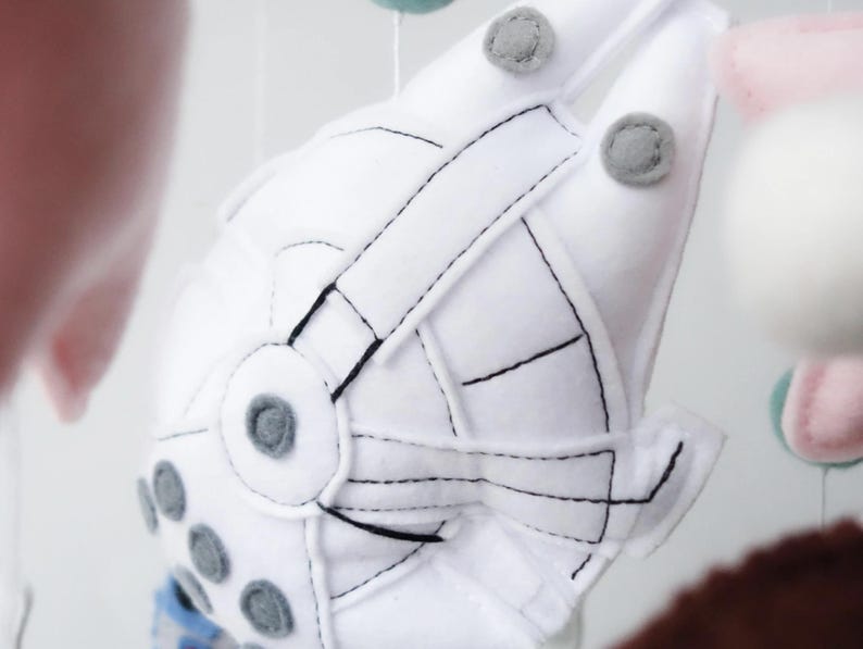 May include: A white felt mobile element shaped like a spaceship, with black stitched details and gray accents. The spaceship has a circular design with gray circles and is suspended with other colorful felt shapes.