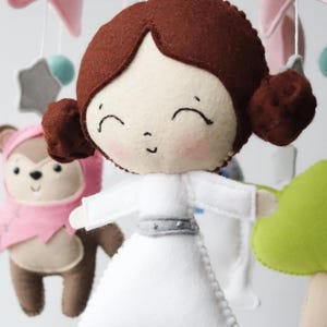 May include: A handmade baby mobile featuring felt figures of a princess in a white dress, a bear in a pink outfit, and other decorative elements. The mobile includes a star, a mushroom, and other shapes in soft colors.
