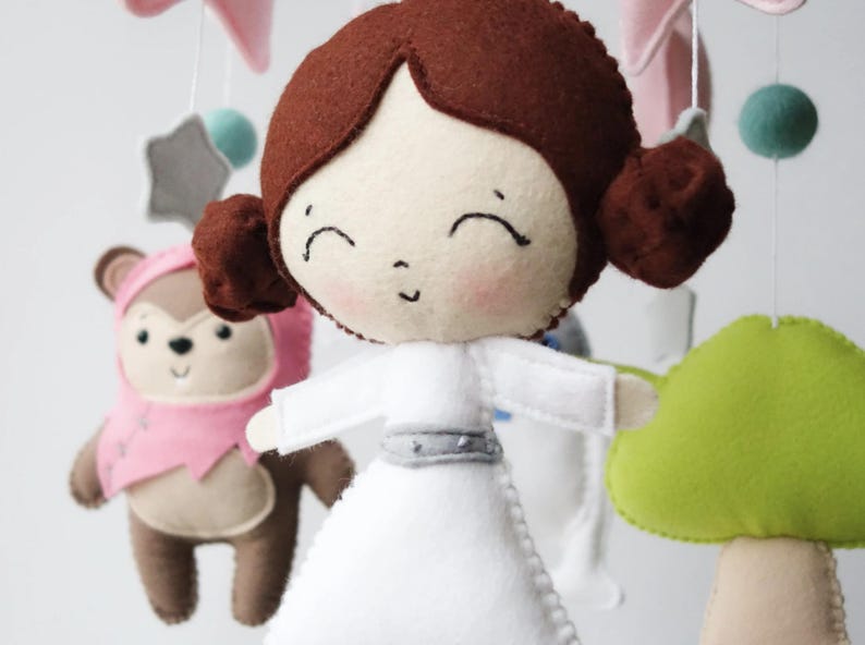 May include: A handmade baby mobile featuring felt figures of a princess in a white dress, a bear in a pink outfit, and other decorative elements. The mobile includes a star, a mushroom, and other shapes in soft colors.