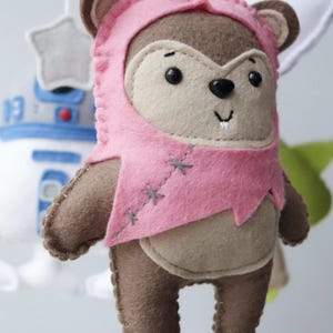 May include: A handmade felt Ewok figure wearing a pink hood and tunic. The plush toy has brown and tan coloring with black eyes and a stitched smile. Other Star Wars themed felt figures are visible in the background.