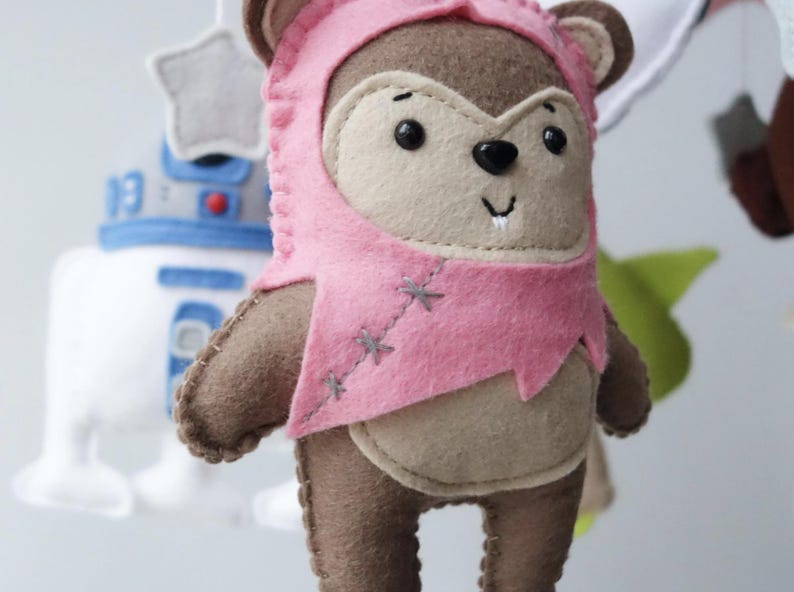 May include: A handmade felt Ewok figure wearing a pink hood and tunic. The plush toy has brown and tan coloring with black eyes and a stitched smile. Other Star Wars themed felt figures are visible in the background.