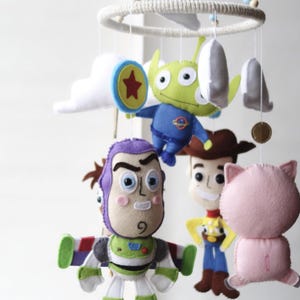 May include: A handmade baby mobile featuring felt characters from the Toy Story movie. The mobile includes Buzz Lightyear, Woody, an alien, Hamm the pig, and other decorative elements. The characters are suspended from a circular frame.