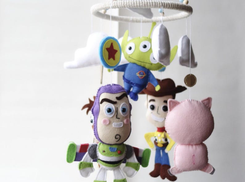 May include: A handmade baby mobile featuring felt characters from the Toy Story movie. The mobile includes Buzz Lightyear, Woody, an alien, Hamm the pig, and other decorative elements. The characters are suspended from a circular frame.