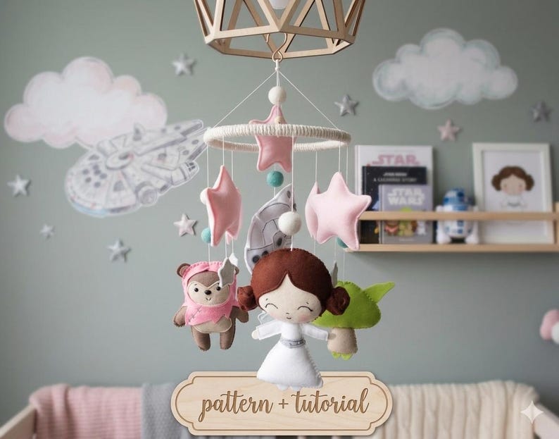 May include: A handmade baby mobile featuring felt Star Wars characters and pink stars. The mobile includes a Princess Leia doll, a Yoda doll, and a Wicket the Ewok doll. The mobile is suspended from a wooden ring and is designed for a nursery.