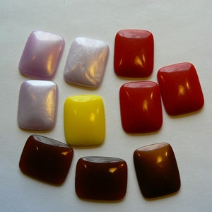 May include: A collection of nine rectangular cabochons with rounded corners, featuring a variety of colors including pearlescent lavender, solid bright yellow, deep red, and dark brown, all with a smooth, polished, and reflective surface finish.