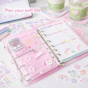 May include: A pink planner with a calendar page open, surrounded by stickers, pens, and a tablet. The planner has a clear cover with decorative shapes. The text "Plan your soft life" is visible.