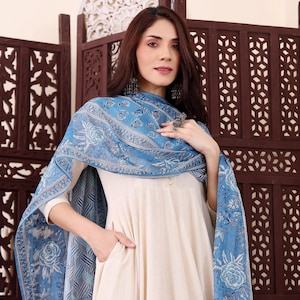 May include: A cream-colored dress with a blue floral patterned scarf. The scarf is draped over the shoulders and features a light blue and white design. The dress has long sleeves and a pocket.
