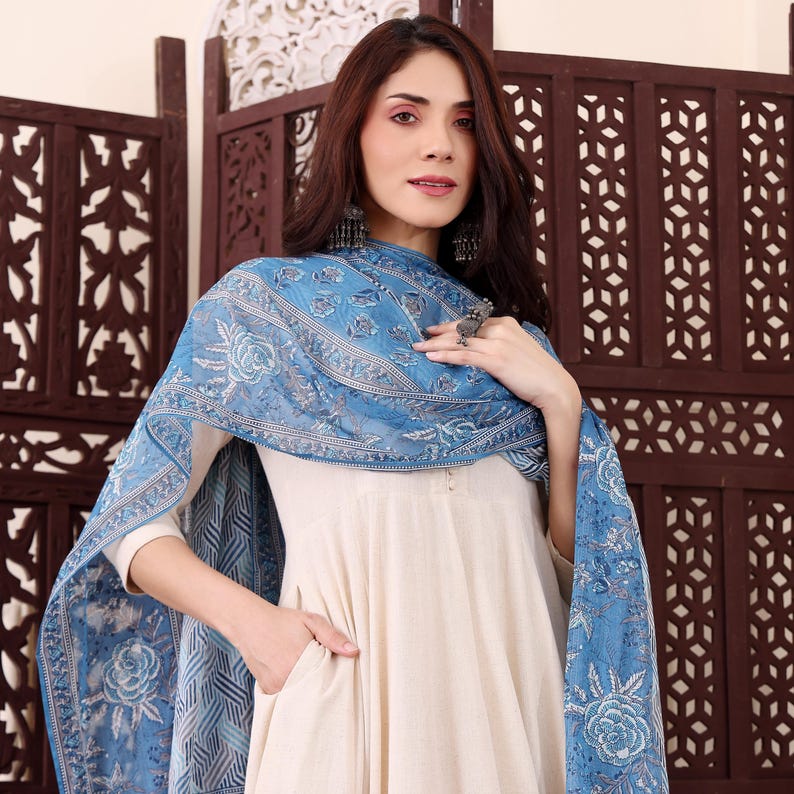 May include: A cream-colored dress with a blue floral patterned scarf. The scarf is draped over the shoulders and features a light blue and white design. The dress has long sleeves and a pocket.